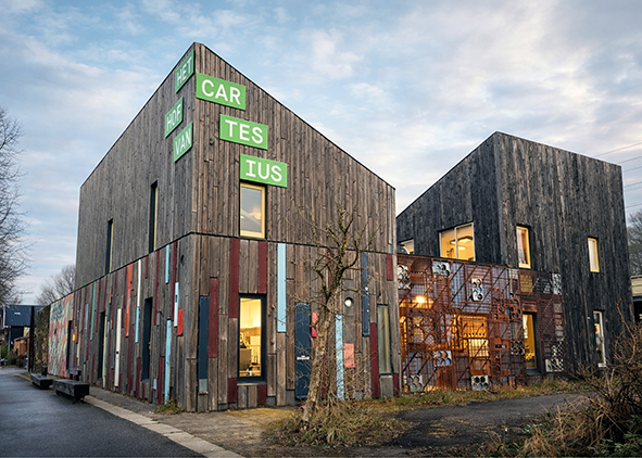 Street-side and corner view of the Brandweer pavilion (gateway building), showing the patchwork façade of reclaimed timber boards and coloured panels, the permeable metal screen and the entrance signature of ‘Het Hof van Cartesius’ (credits: the Authors, 2025).  AGATHÓN 19 | 2026